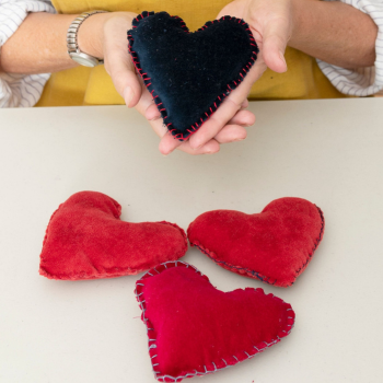 Person holding a handmade black heart-shaped cushion, with three red fabric hearts on the table, all stitched with visible thread.
